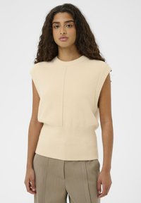 Eine Frau mit langen, lockigen Haaren, die einen ärmellosen beigen Pullover und taubfarbene, hochgeschnittene maßgeschneiderte Hosen trägt und vor einem schlichten Hintergrund steht.