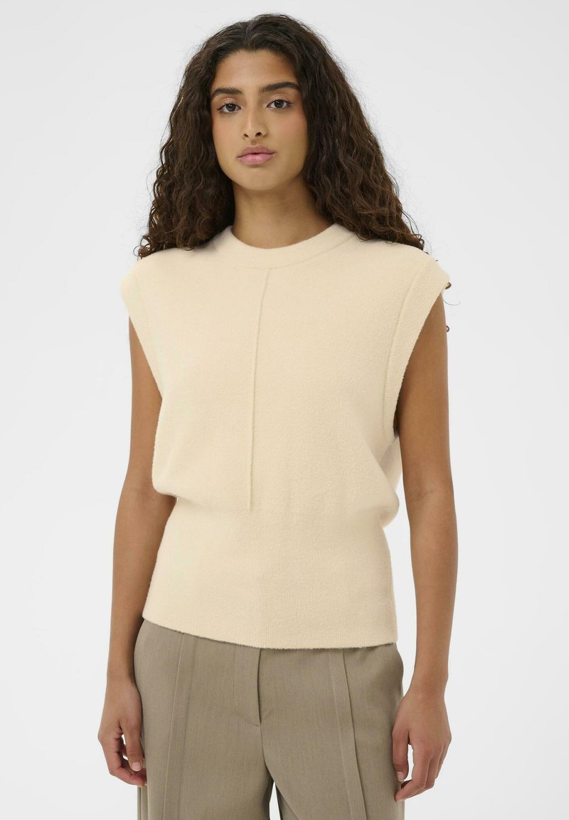 Eine Frau mit langen, lockigen Haaren, die einen ärmellosen beigen Pullover und taubfarbene, hochgeschnittene maßgeschneiderte Hosen trägt und vor einem schlichten Hintergrund steht.