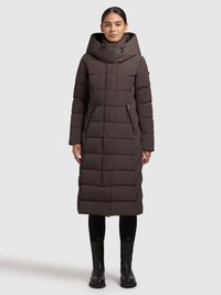 Pitkä, pullea ruskea takki, jossa on korkea kaulus ja iso huppu. Siinä on tikattu malli, kaksi vetoketjulla suljettavaa sivutaskua ja se on yhdistetty mustien saappaiden kanssa.