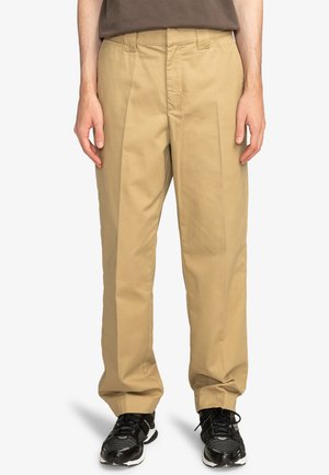 Beige cotton trousers with a straight leg design, pleated front, and side pockets. Worn with black sneakers featuring mesh accents.