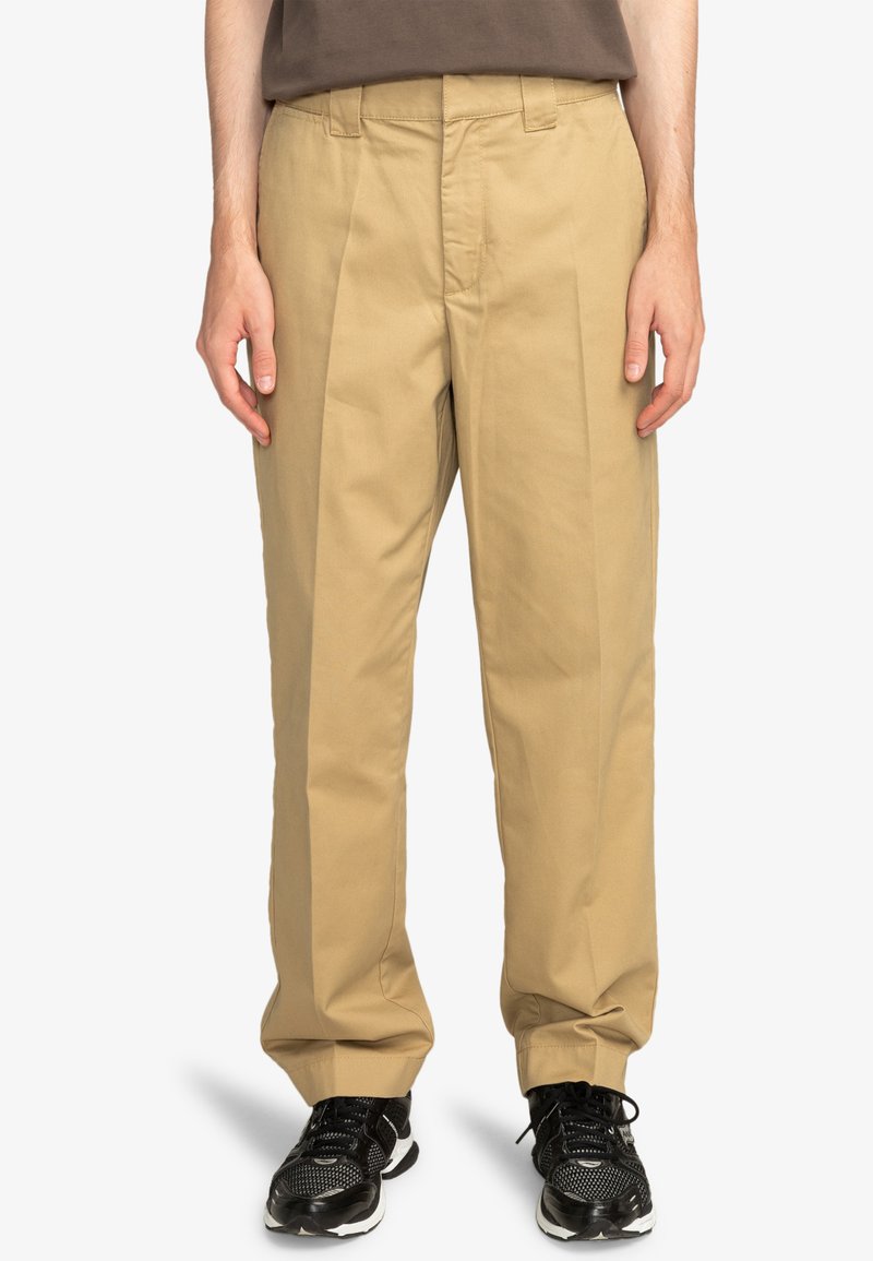 Beige bomuldsbukser med lige ben, plisseret front og sidelommer. Bæres med sorte sneakers med mesh-detaljer.