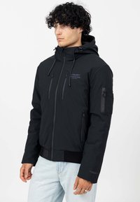 Zwarte zip-up jas met een capuchon, meerdere ritszakken en geribde manchetten. Gemaakt van gladde stof met een logo op de borst en de mouw.