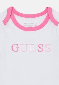 Body en coton biologique blanc avec un col en bord côte rose. Présente "GUESS" brodé en lettres roses dégradées. Taille 12M.