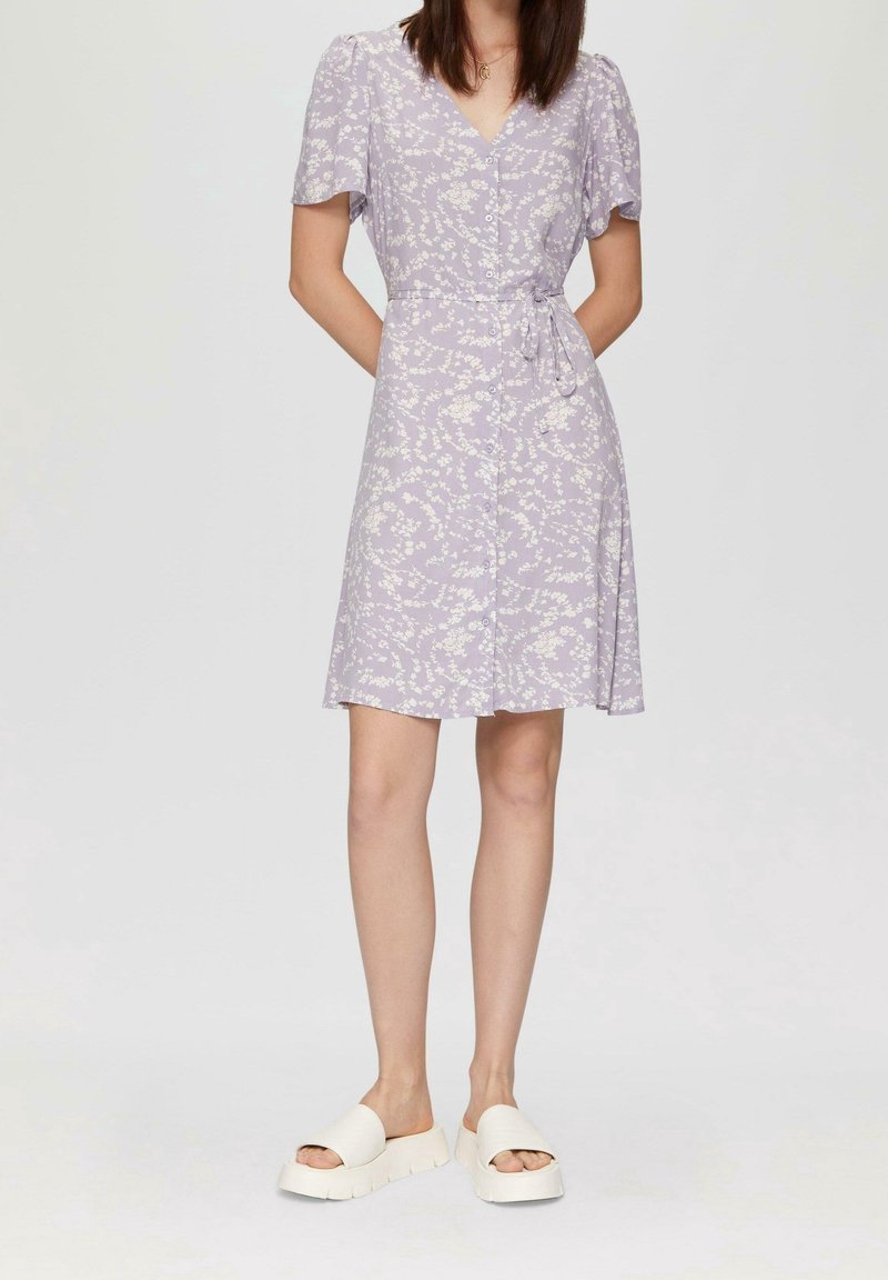 Femme portant une robe midi lavande à fleurs avec des boutons et une ceinture à la taille, associée à des sandales blanches à plateformes.