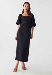 Black midi dress with puff sleeves, textured horizontal stripes at the bodice, and a straight skirt. Worn with nude heeled sandals.