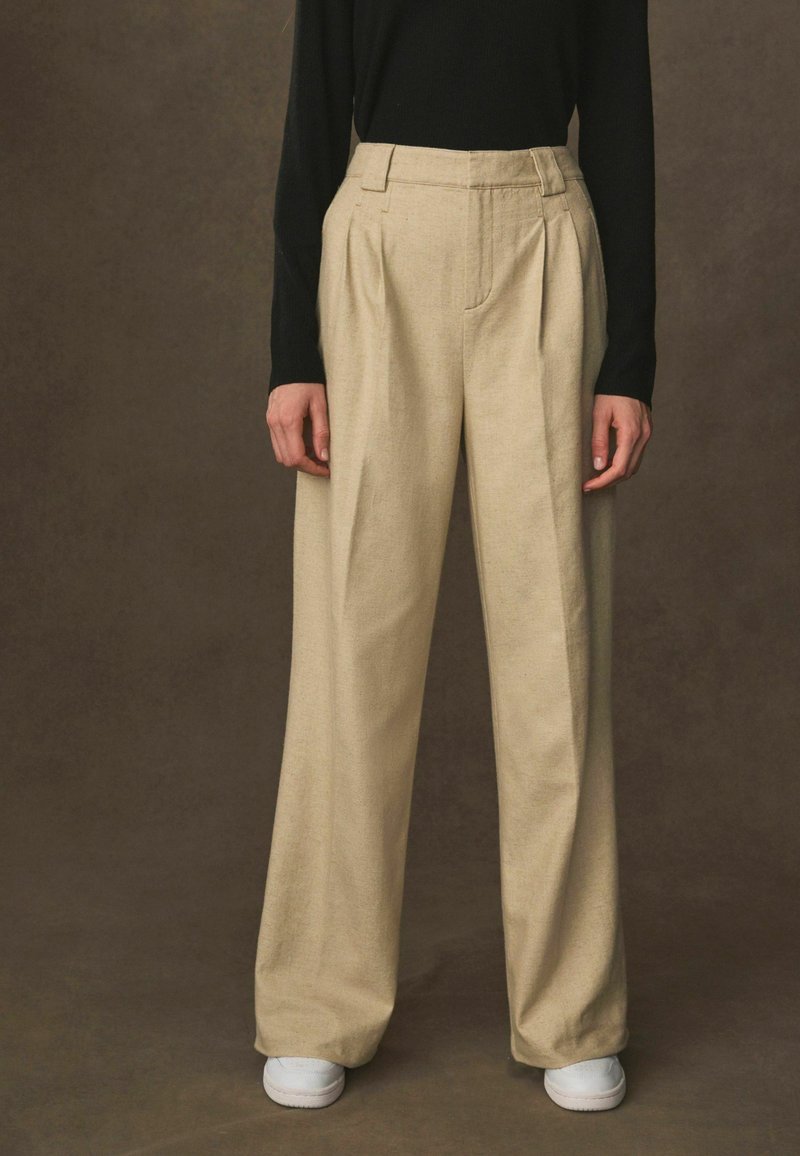 Beige, plisserede bukser med vidde, lavet af et tekstureret stof, med høj talje og sidelommer, parret med hvide sneakers.