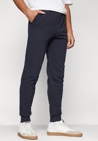Pantaloni della tuta navy con cintura elastica, tasche laterali e caviglie elastiche. Tessuto misto cotone con una superficie liscia. Abbinati a sneaker bianche.