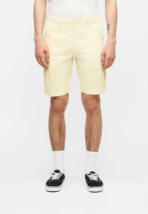 Férfi bézs színű, térdig érő cargo rövidnadrágban, fehér zokniban és fekete tornacipőben, karjait lazán oldalán tartva áll.