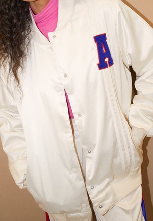 Personne portant une veste varsity en satin blanc avec un écusson lettre A bleu et rouge, associée à un col roulé rose vif en dessous.