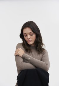 Bodysuit en tela marrón claro con mangas largas, que presenta un cuello alto y un acento plateado en la muñeca. Tela negra en la mitad inferior.