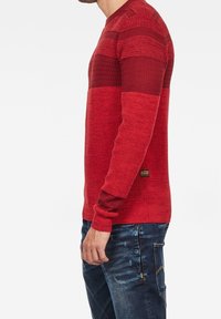 Maglione rosso lavorato a maglia con strisce testurizzate e design morbido. Il materiale sembra morbido con una forma aderente. Include un'etichetta del marchio sul lato.