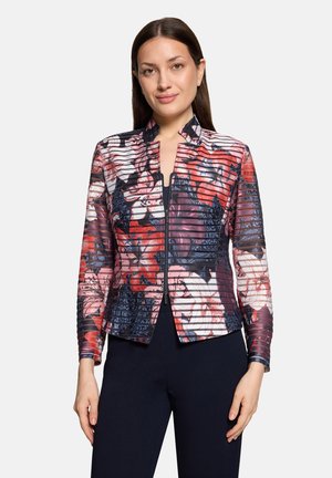 Woman with straight dark hair wearing a fitted zip-up jacket with red, pink, white, and navy floral patterns, standing against a plain background.