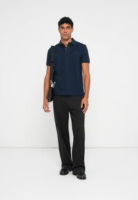 Camisa polo azul-marinho de mangas curtas, com gola e riscas verdes em contraste; combinada com calças largas pretas e sapatos pretos.