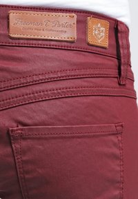 Bordeauxfärgade byxor med synlig sömnad, en läderetikett på midjebandet med "Freeman T. Porter" och en liten emblem. Slät textur.