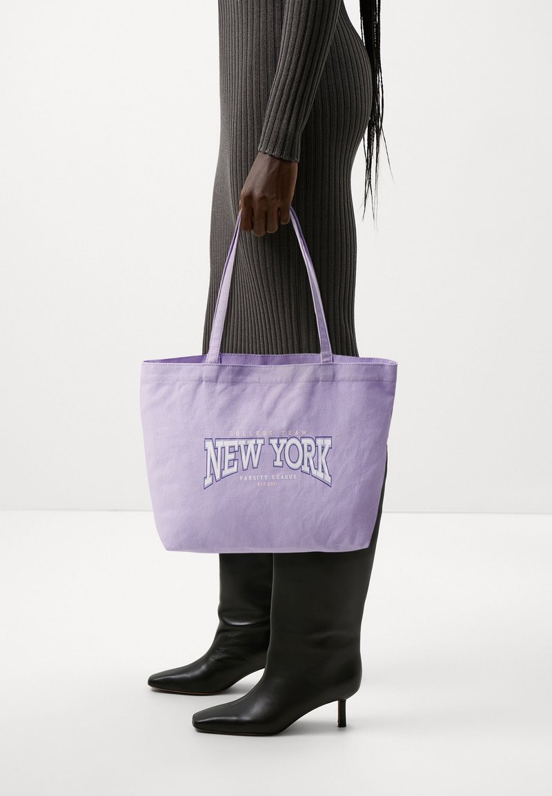 Lawendowa torba na ramię z tkaniny z napisem "NEW YORK" w białym i fioletowym kolorze, dwoma materiałowymi uchwytami, prostokątnym kształtem oraz gładką teksturą.