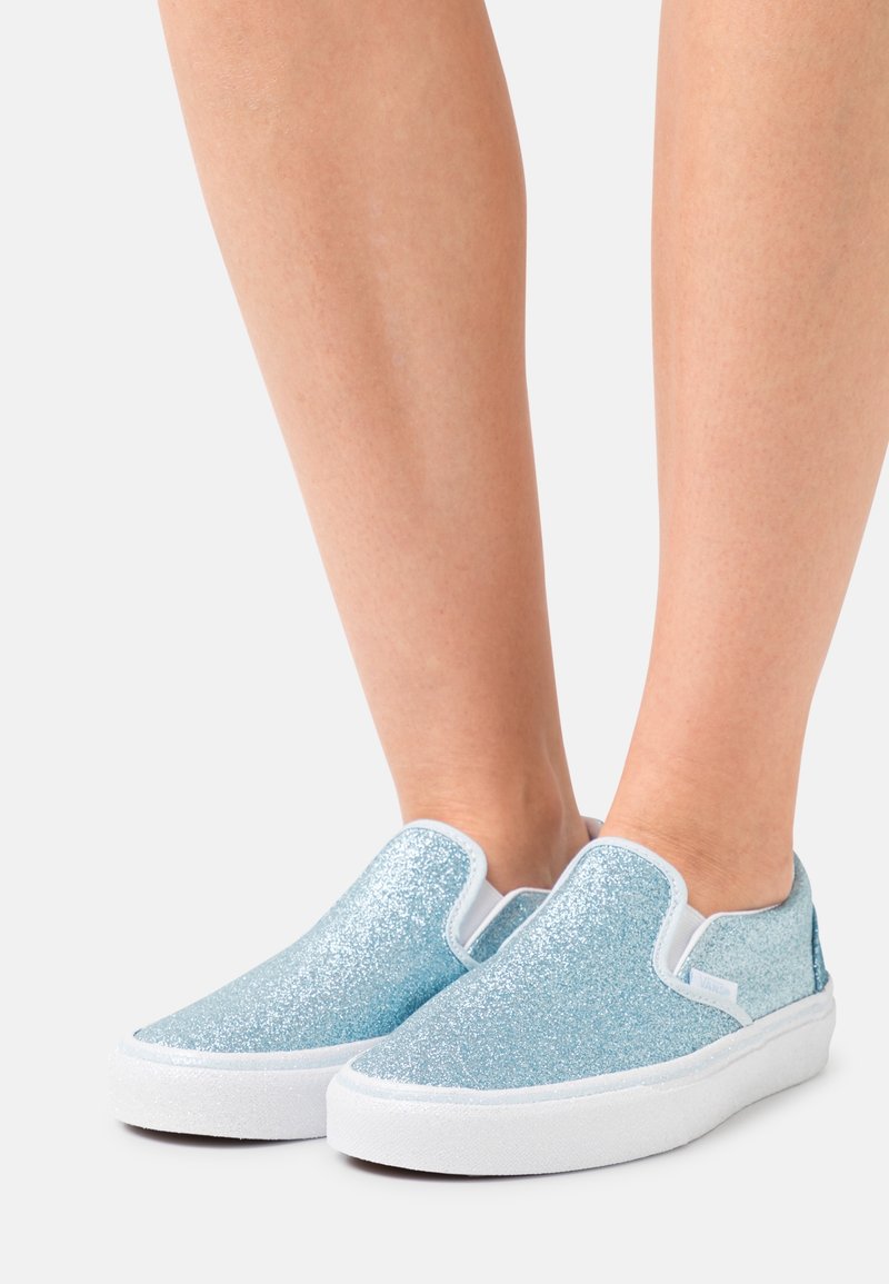 Personne portant des baskets slip-on bleu clair à paillettes avec des semelles blanches, debout sur fond blanc. Seules les jambes inférieures et les pieds sont visibles.