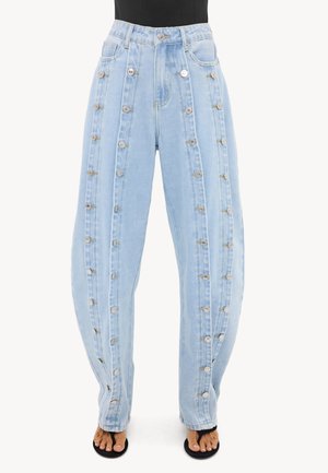 Jean taille haute bleu clair avec des doubles rangées verticales de boutons argentés sur le devant et les coutures latérales, porté avec un haut noir et des sandales noires ouvertes à l’avant.