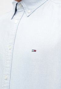 Jasnoniebieska koszula z guzikami, wykonana z bawełny, z małym haftowanym logo w czerwonych i granatowych akcentach na klatce piersiowej. Gładka faktura.