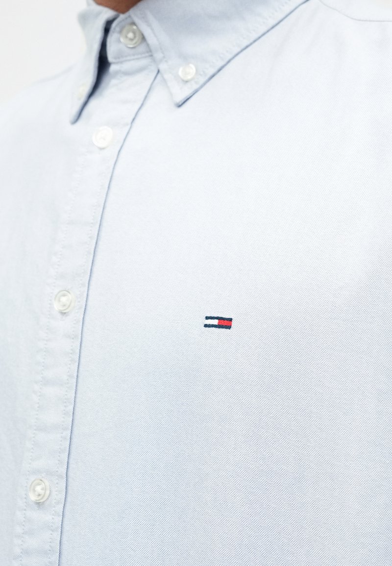 Light blue button-up shirt made of cotton, featuring a small embroidered logo with red and navy accents on the chest. Smooth texture.