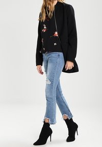 Abrigo largo negro sobre una blusa negra con estampado floral, combinado con jeans ajustados desgastados en azul claro y botas de tobillo negras de punta.