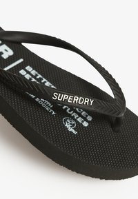 Tongs noires avec un pied en caoutchouc texturé, fines sangles noires et un logo "SUPERDRY" argenté sur la sangle, présentant un motif en chevrons.