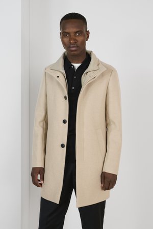 Abrigo de mezcla de lana beige con un cuello alto, botones visibles y bolsillos laterales, llevado sobre una camiseta de polo negra, contra un fondo neutro.