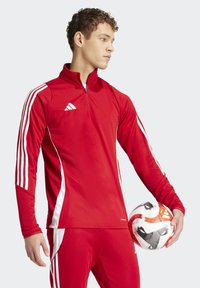 Red athletic zip-up jacket with white stripes on sleeves, made of smooth fabric. Model holds a patterned soccer ball. Features Adidas logo.