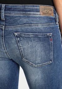 Pantalones vaqueros de mezclilla azul oscuro con un diseño de tiro medio, sutiles desgastes y un parche de cuero en la parte posterior de la cintura con detalles de la marca.