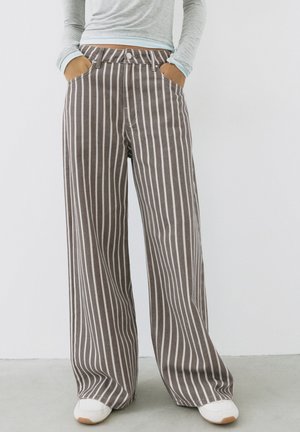 Person iført brune vidde bukser med hvide lodrette striber, hænderne i lommerne, kombineret med hvide sneakers og en lysegrå langærmet top.