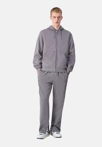 Sivá mikina na zips s klokaniimi vreckami a zodpovedajúcimi teplákmi, ktoré majú uvoľnený strih a mäkkú látku. Biele tenisky dokončujú outfit.