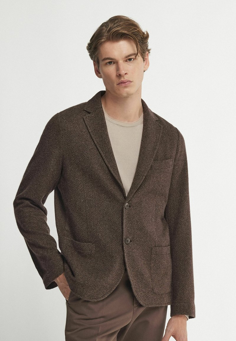 Brauner Hahnentrittmuster-Blazer mit zwei Vorderaschen, Reverskragen und zwei Knöpfen, getragen über einem beigen Hemd aus strukturiertem Wollmaterial.