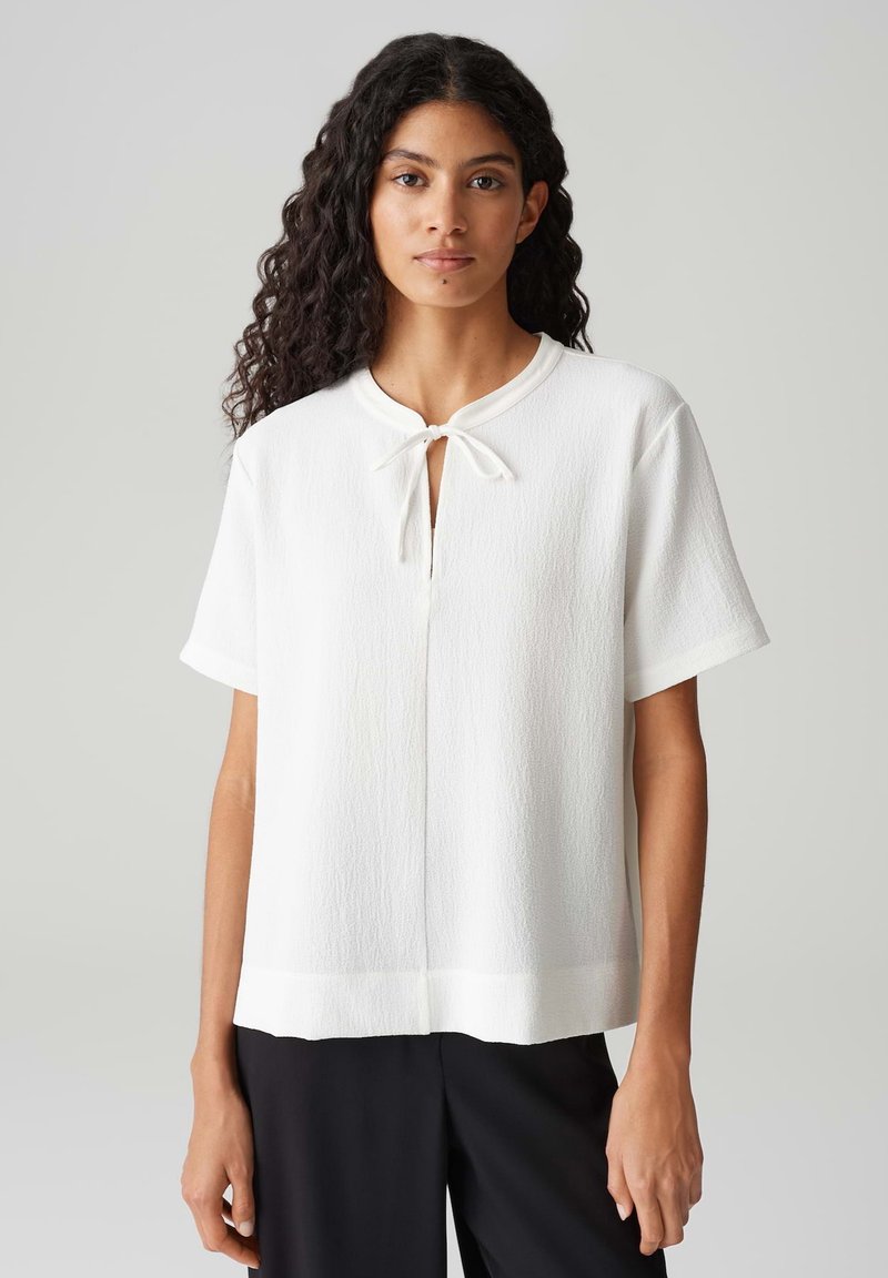 Haut blanc à manches courtes avec un col rond et un détail en trou de serrure noué avec un nœud. Tissu texturé, coupe ample et ourlet droit.