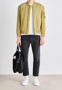 Bomber bunda olivově zelená, bílé tričko, černé džíny, bílé tenisky, držící černou tašku. Hladká textura, přiléhavý střih, minimalistické detaily.