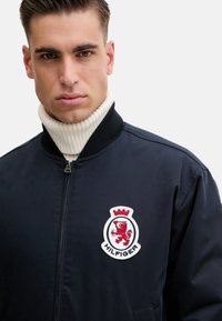 Chaqueta bomber de la marina con cuello acanalado, featuring un logo bordado en rojo y blanco en el pecho y una textura suave y brillante.