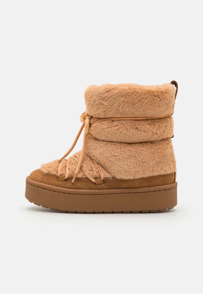 Bottines en fausse fourrure couleur tan avec une semelle épaisse et texturée. Elles présentent un haut replié, un détail noué et une base en suede lisse avec une semelle légère.
