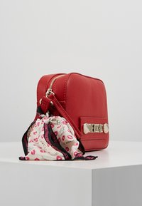 Punainen tekonahkainen käsilaukku suorakulmaisella muotoilulla. Varustettu kullanvärisellä metallisoljella ja koristeellisella huivilla, jossa on huulipainatuksia.