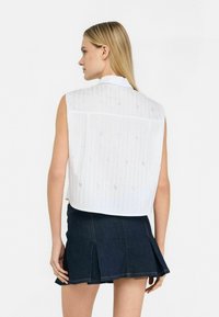 Camicetta bianca senza maniche con linee verticali testurizzate e colletto, abbinata a una gonna a pieghe in denim scuro. La camicetta presenta dettagli delicati.