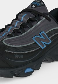 Κοντινή λήψη μαύρου και γκρι New Balance 1000 sneaker με μπλε λεπτομέρειες, γλώσσα με κυματιστό μοτίβο και μεγάλο λογότυπο "N" στο πλάι.