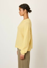 Rumeno, puhasto pulover z sproščenim krojem, okroglim izrezom in dolgimi rokavi. Nošen čez teksturirane, bež, stranske hlače.