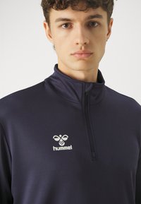 Jonge man met kort krullend haar die een marineblauwe sportjas met ritssluiting draagt met een wit hummel-logo op de borst.