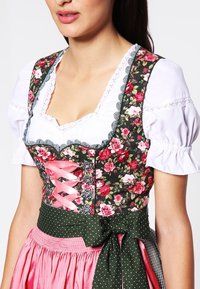 Dirndl jurk met bloemmotief, kenmerkend door een aansluitend lijfje met roze linten, witte pofmouwen en een groene schort met stippen, gecombineerd met een roze rok.