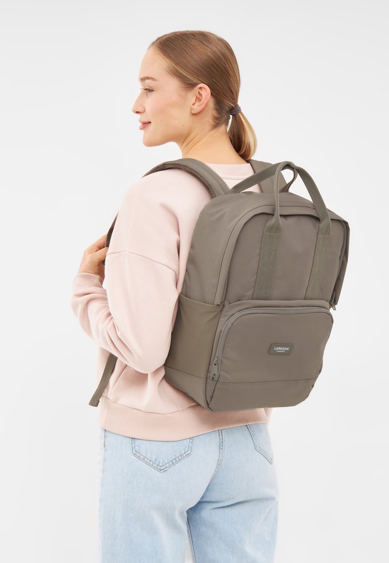 Zaino grigio realizzato in tessuto morbido con un design strutturato, dotato di due manici superiori, spallacci imbottiti e una tasca anteriore con zipper.