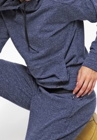 Survêtement de sport bleu marine en tissu texturé chiné. Comprend une capuche avec cordon de serrage et une poche zippée sur le pantalon.