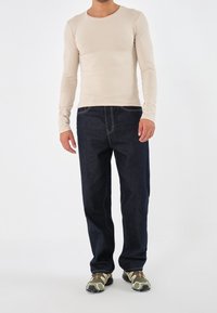 Beige långärmad skjorta, mörkblå denimjeans, sneakers med kamouflagemönster, avslappnad passform med kontrasterande sömdetaljer på jeansen.