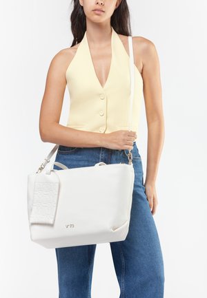 Donna con top giallo senza maniche e jeans blu che tiene una grande borsa bianca con targhetta in rilievo e ferrature dorate.