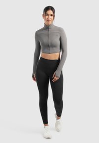Grau zugeschnittene Zip-Jacke mit langen Ärmeln, strukturierten Panels und Daumenlöchern, kombiniert mit hochtaillierten schwarzen Leggings und weißen Turnschuhen.