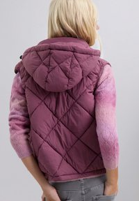 Persona con cabello rubio que lleva un chaleco acolchado con capucha morado sobre un suéter ombré rosa y pantalones grises, de pie contra un fondo liso.