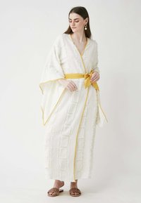 Long white kimono with textured patterns, yellow trim, and a tied waist. Pair of brown slip-on sandals complements the outfit.