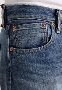 Blå denimjeans med en närbild av en framficka, med kopparnitar, kontrasterande sömmar och en texturerad yta.