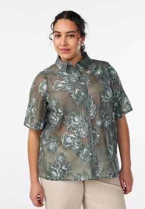 Naine, kes kannab läbipaistvast rohelisest lühikeste varrukatega pluusist valgete lillemustritega ja beeže pükse, seisab lihtsa tausta ees.